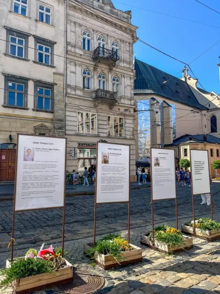 Four memorial stands with portraits and text on a cobblestone street in Lviv, with historic buildings and a church in the ...
