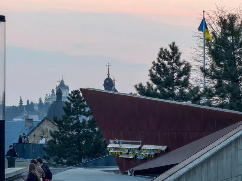 Lviv skyline with church domes, a modern building, people on a path, and a Ukrainian flag at dusk.