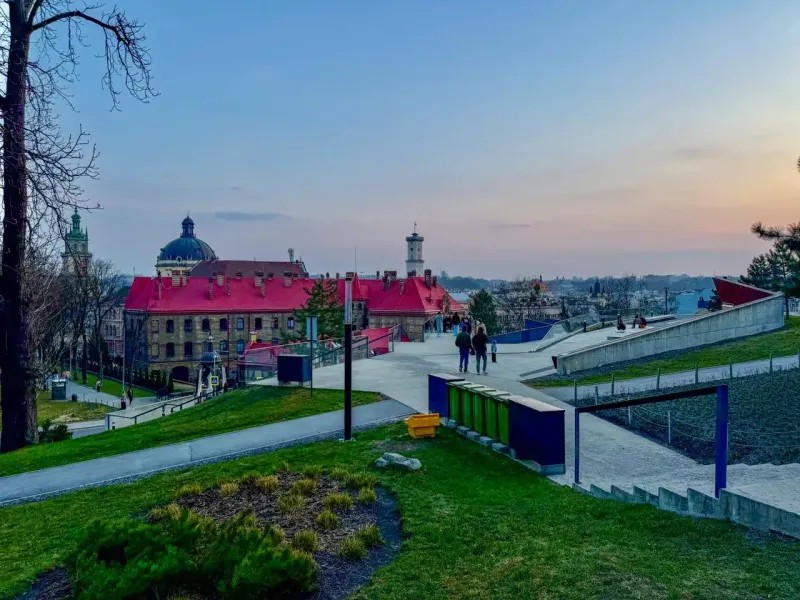 View from High Castle Hill in Lviv at sunset, showing red-roofed buildings, a dome, a tower, and modern park paths with pe...