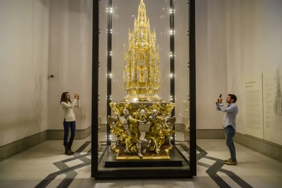 Una custodia ornamentada y dorada en una vitrina de cristal, fotografiada por dos visitantes con sus teléfonos.