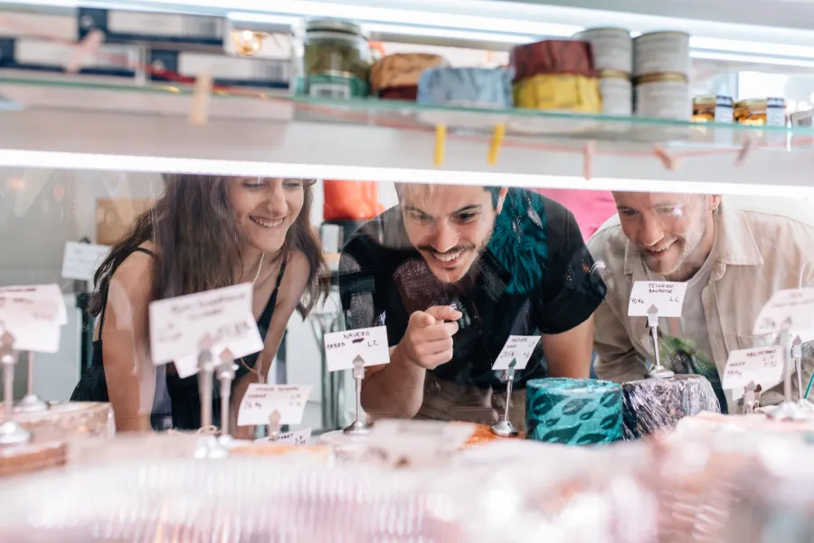 Tres jóvenes sonríen mientras miran una selección de quesos a través de un mostrador de cristal en una tienda.