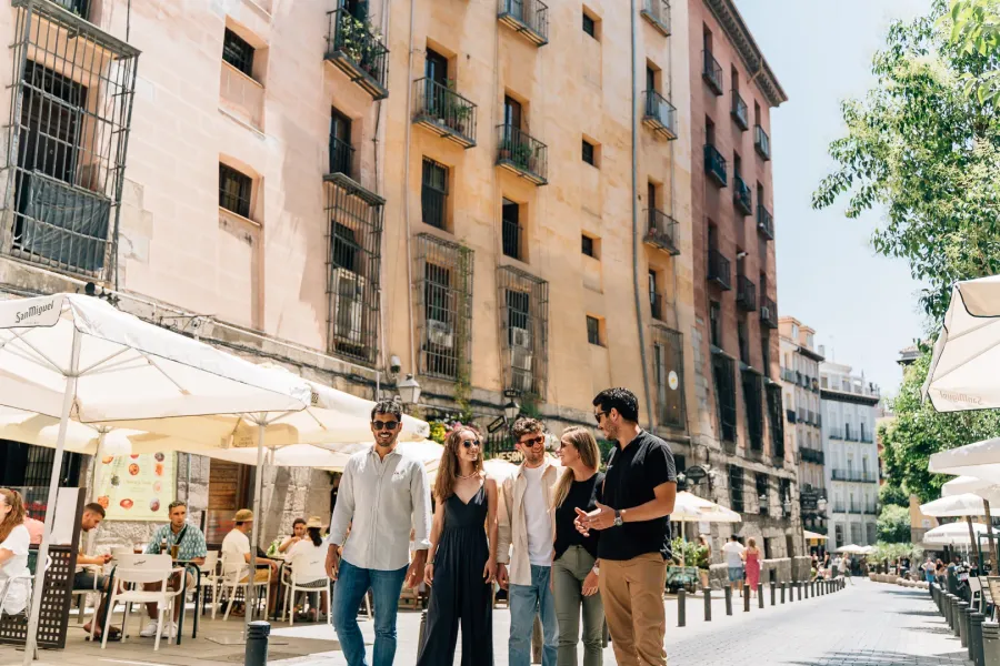 Un grupo de cinco jóvenes camina por una soleada calle de Madrid bordeada de mesas de café y edificios históricos.
