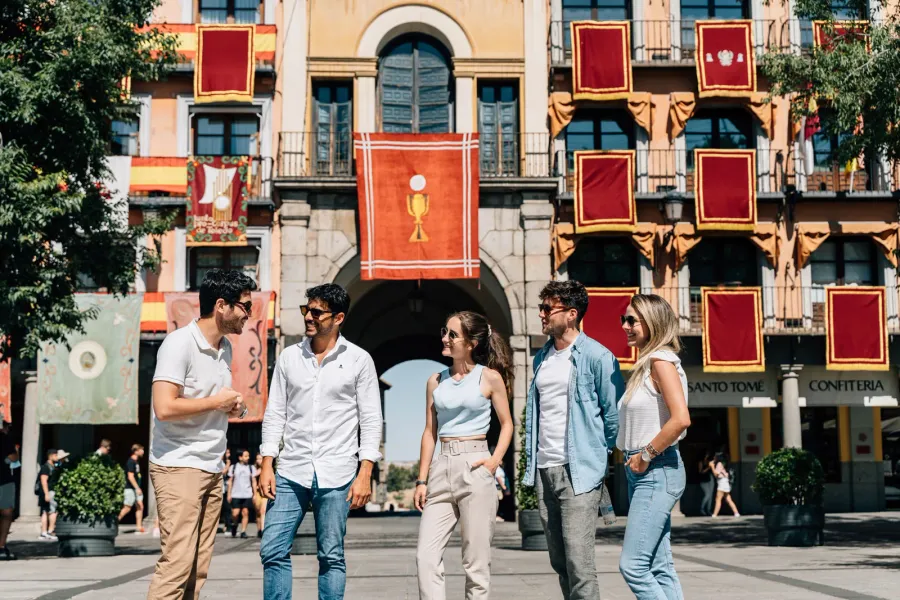 Un grupo de cinco jóvenes en una plaza soleada, frente a un edificio decorado con estandartes rojos y dorados.