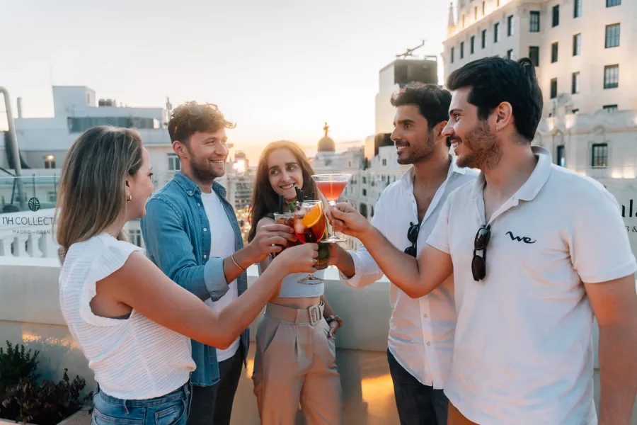 Cinco jóvenes amigos brindan con cócteles de colores en una azotea de Madrid al atardecer.