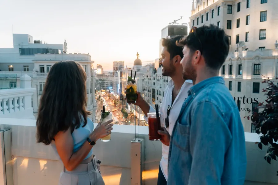 Tres amigos con cócteles en una azotea con vistas a las concurridas calles de Madrid al atardecer.