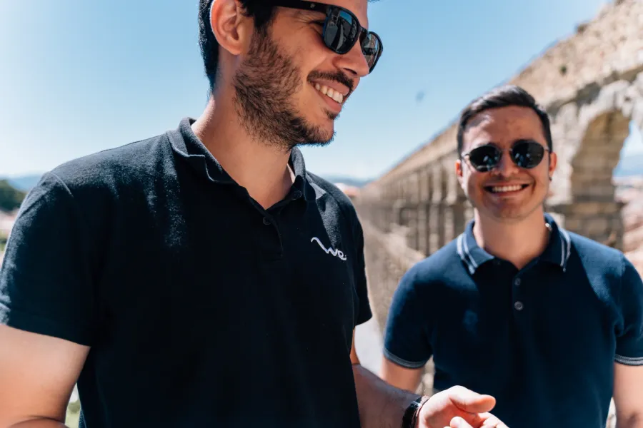 Dos jóvenes con gafas de sol sonríen frente al Acueducto de Segovia en un día soleado.