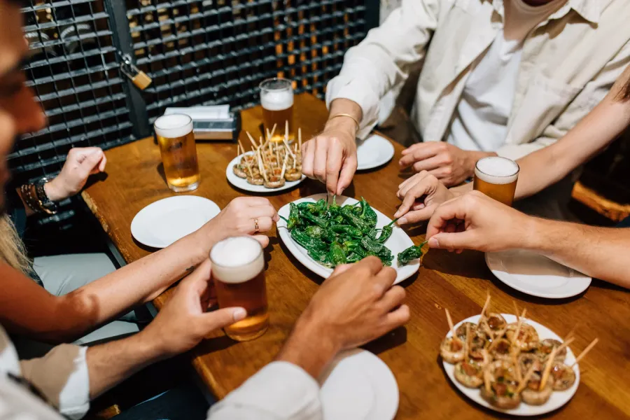 Amigos compartiendo tapas, incluyendo pimientos de Padrón, y bebiendo cerveza en una mesa de madera en un bar.