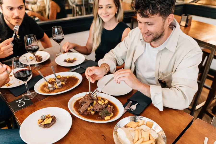 Tres personas en una mesa de madera compartiendo platos de estofado de ternera y bebiendo vino tinto en un restaurante.