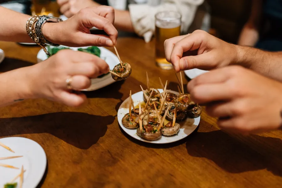 Manos cogiendo tapas de champiñones con palillos de un plato en una mesa de madera.