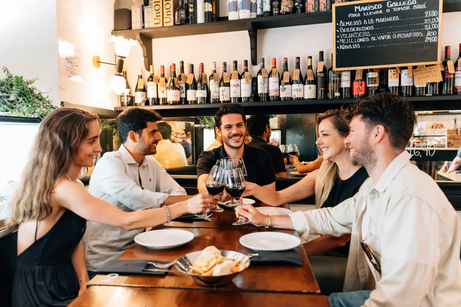 Un grupo de cinco amigos sentados a una mesa en un bar, sonriendo y brindando con copas de vino tinto.