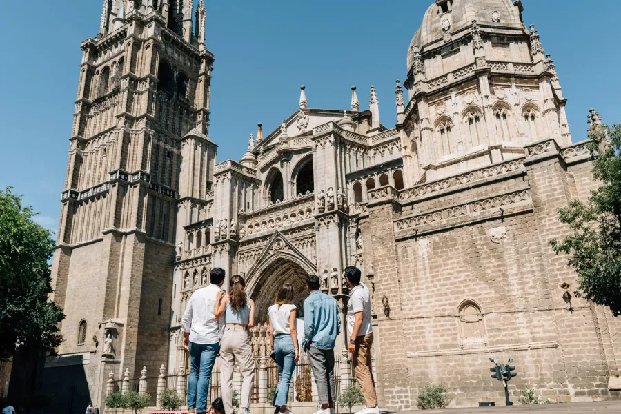 Un grupo de cinco personas, vistas desde atrás, admiran la intrincada fachada de piedra de la Catedral de Toledo en un día...