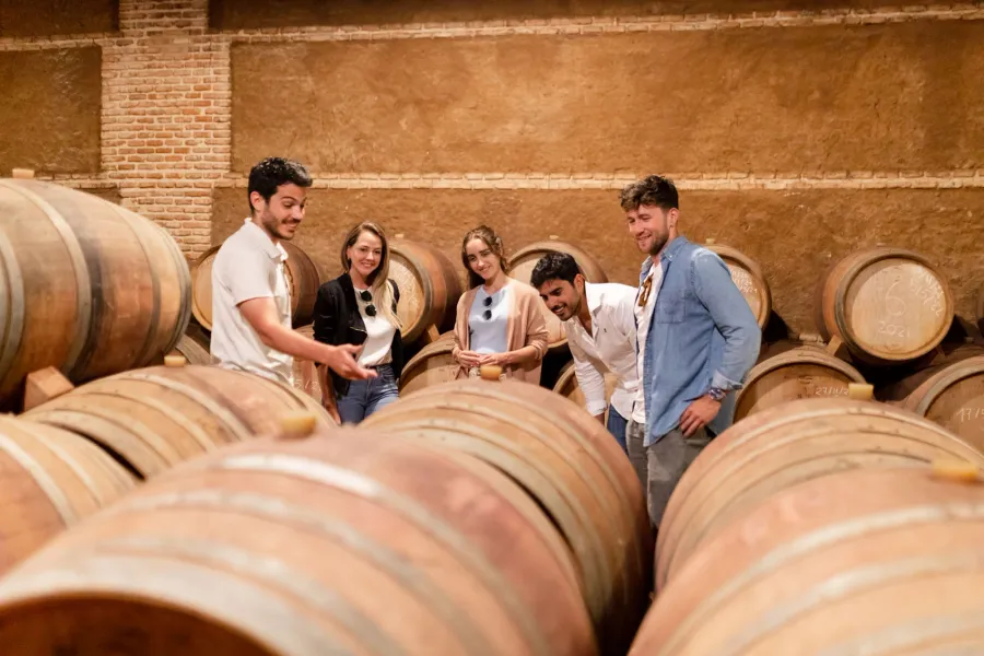 Un grupo de turistas en una bodega, rodeados de docenas de barriles de madera apilados contra las paredes.