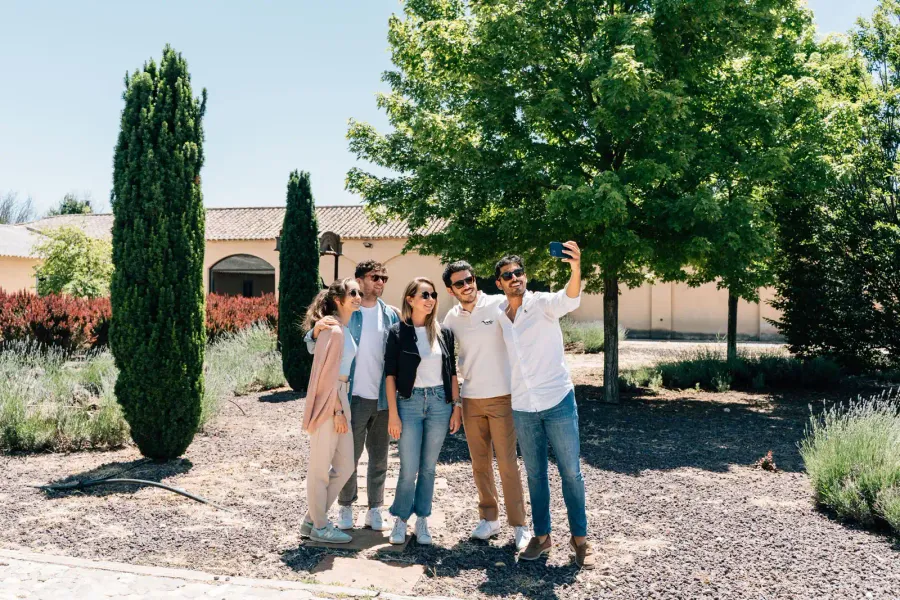 Un grupo de cinco amigos se hace un selfi sonriente en un jardín soleado con cipreses y una villa al fondo.