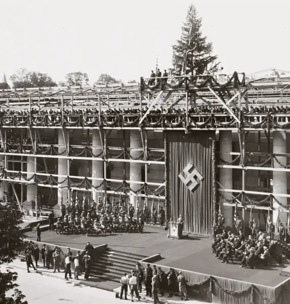 Schwarz-weiß-Foto eines im Bau befindlichen Gebäudes, geschmückt mit Girlanden und einem großen Hakenkreuzbanner. Menschen...