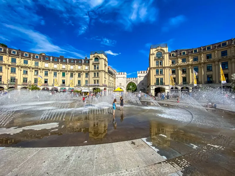 People enjoying the large fountain at Karlsplatz in Munich, with Karlstor archway and historic buildings in the background...