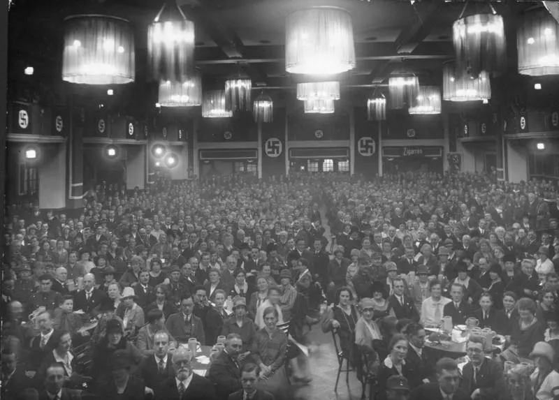 Interior of a crowded Munich beer hall during the Nazi era with swastika banners and chandeliers.