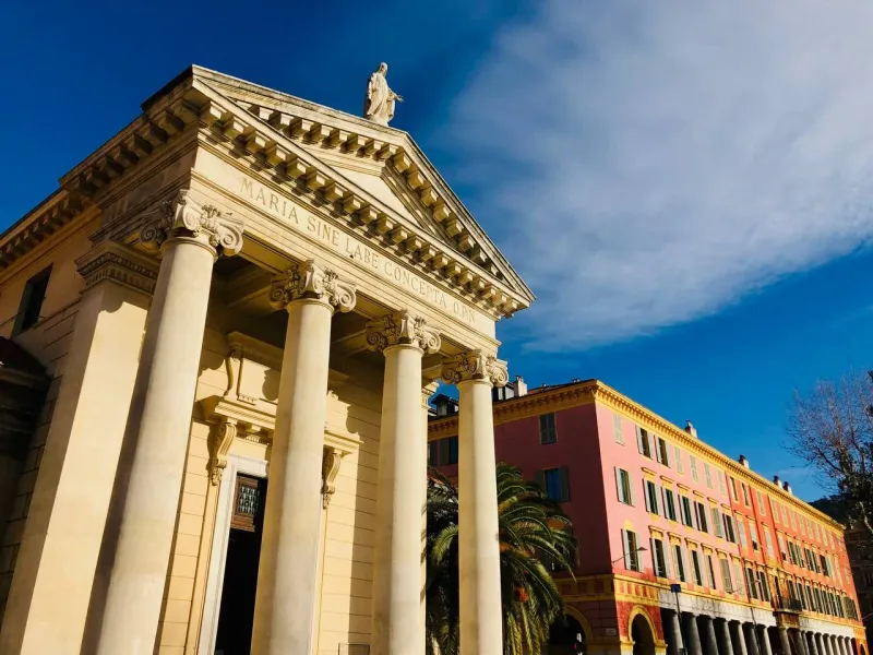 Neo-classical chapel with columns, pediment, and statue under a blue sky, next to a pink building with arcades in Nice.
