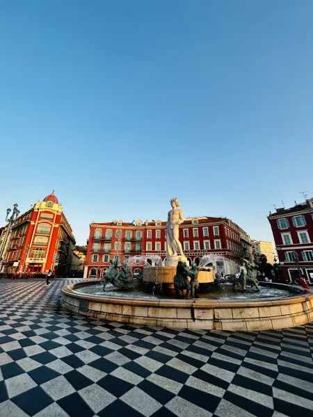Fontanna Słońca (Fontaine du Soleil) w Nicei ze statuą Apolla i brązowymi postaciami, otoczona czerwonymi budynkami pod bł...