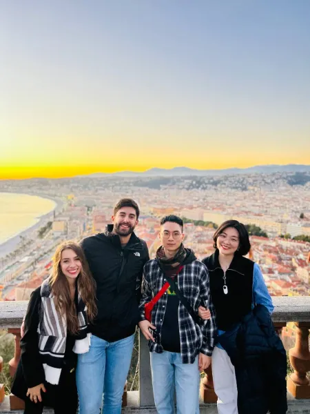 Four people posing on a viewpoint overlooking Nice city, the bay, and mountains at sunset. Golden light illuminates the ho...