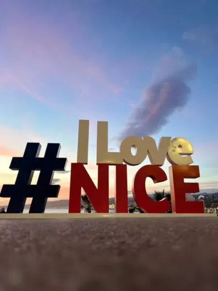 Duży znak „#ILoveNICE” podświetlony o zmierzchu, z różowo-niebieskim niebem nad miastem i morzem w Nicei.