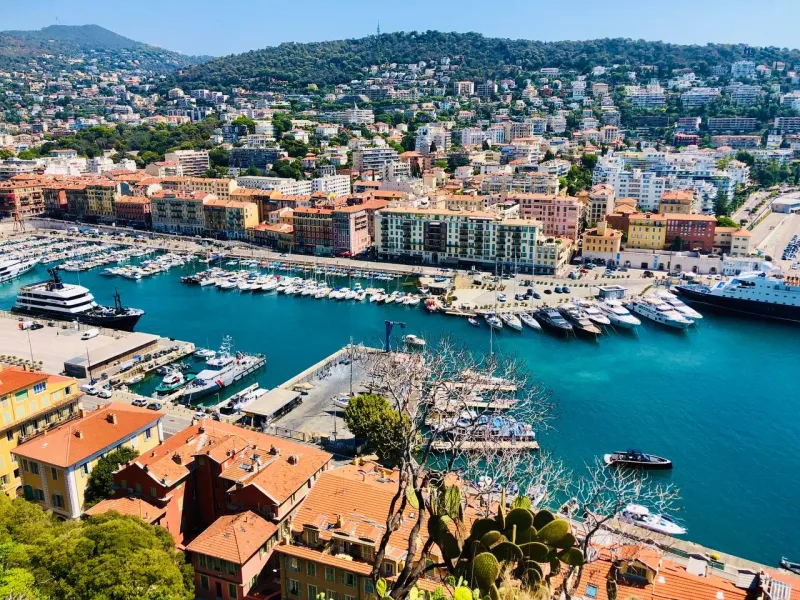 Panoramic view of Nice's Port Lympia with numerous yachts, colorful waterfront buildings, and green hills under a clear bl...