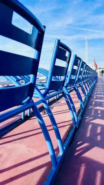 A row of iconic blue chairs on the Promenade des Anglais in Nice, facing the sea under a clear blue sky. Shadows extend on...