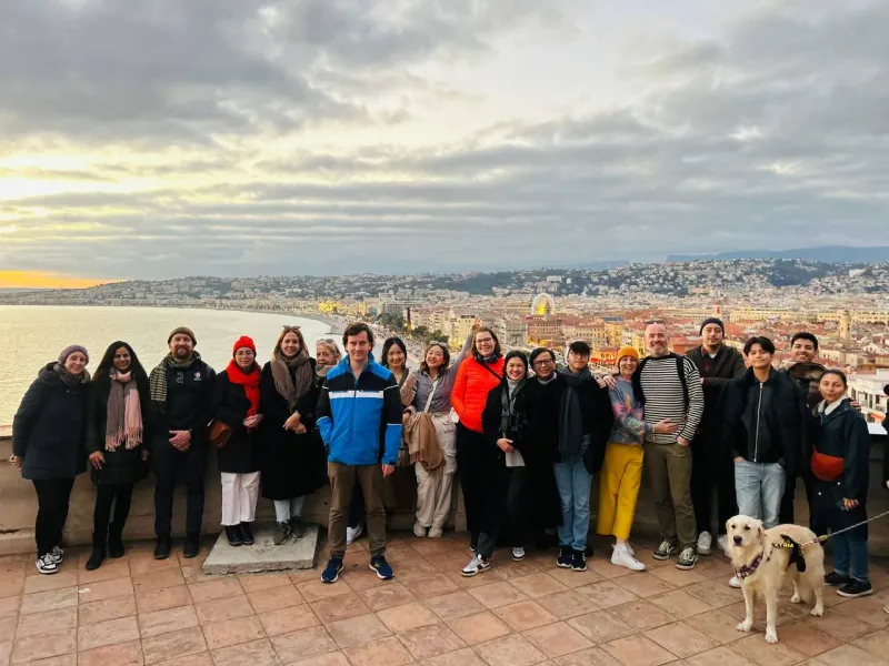 Diverse group and a golden retriever dog on a tiled terrace overlooking Nice city, bay, and coastline at sunset.