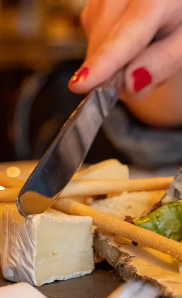 Primer plano de una mano con esmalte rojo cortando un trozo de queso de corteza blanca en un plato con palitos de pan.
