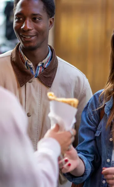 Smiling man watching a crepe being handed to a person in a denim jacket on a street.