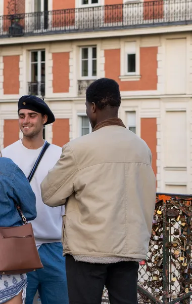Two people, one smiling in a beret next to another from behind, with love locks and a building in background.