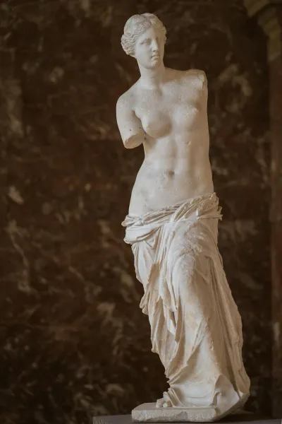 Estatua de mármol blanco de la Venus de Milo sin brazos, parcialmente cubierta, sobre una pared de mármol marrón oscuro.