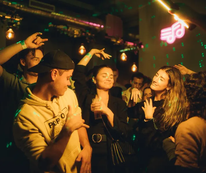Menschen tanzen in einem schwach beleuchteten Club oder einer Bar, mit grünen Laserlichtern und einem pinken Neon-Musiknot...