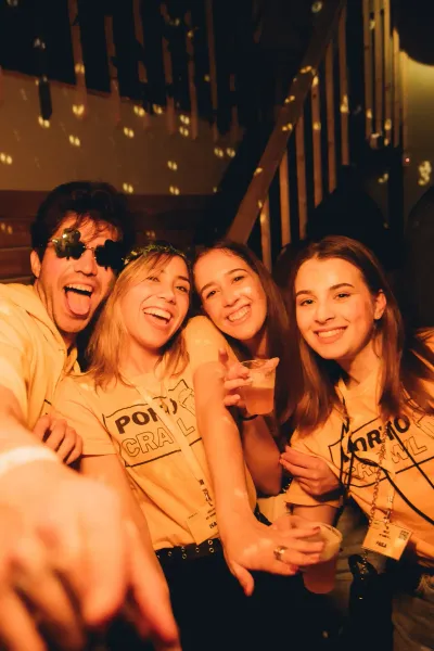 Vier junge Leute, zwei Männer und zwei Frauen, lächeln in einer Bar. Sie tragen gelbe 'PORTO PUB CRAWL' T-Shirts und halte...