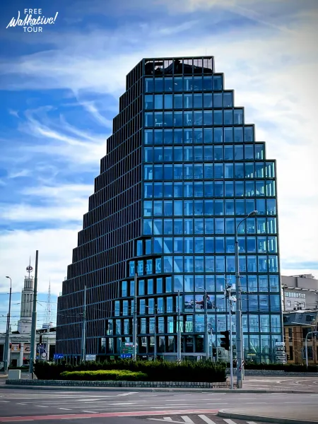 Moderno edificio Bałtyk en Poznań con fachada de vidrio escalonada, bajo cielo azul, con calle y torre histórica visibles.