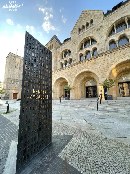 Dunkles Denkmal mit goldenen Zahlen und 'HENRYK ZYGALSKI' auf einem gepflasterten Platz, vor dem Posener Kaiserschloss mit...