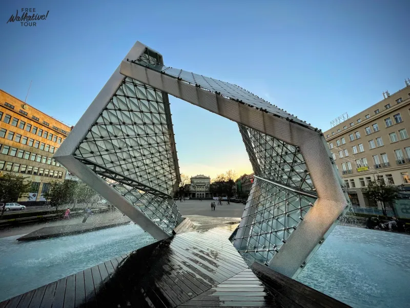 Fuente moderna de vidrio y metal con chorros de agua, en forma de cubo abierto, en una plaza de la ciudad al anochecer. Ed...