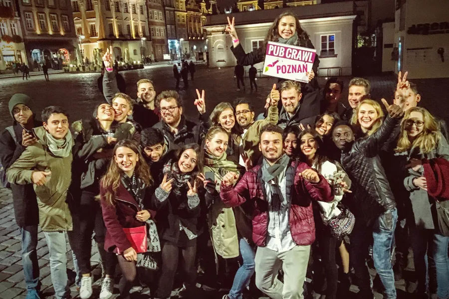 Large group of smiling young adults on a wet cobblestone street at night, one holding a 'PUB CRAWL POZNAN' sign, historic ...