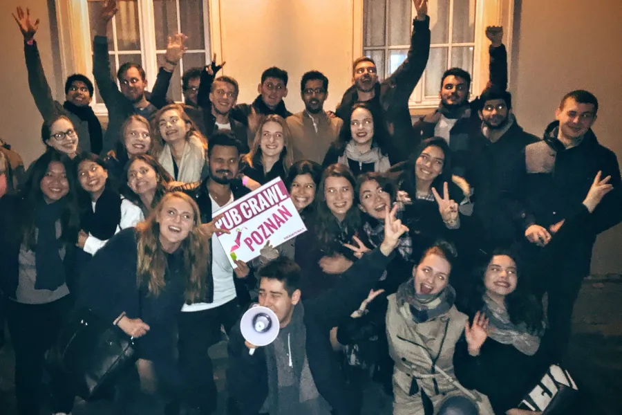 A group of young adults smiling and waving on a pub crawl, holding a "PUB CRAWL POZNAN" sign. One person holds a megaphone.
