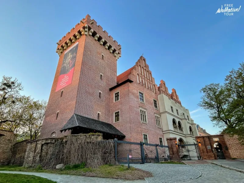 Widok na Zamek Królewski w Poznaniu, ceglany budynek z wysoką wieżą, szczytową fasadą i starym murem, pod błękitnym niebem.