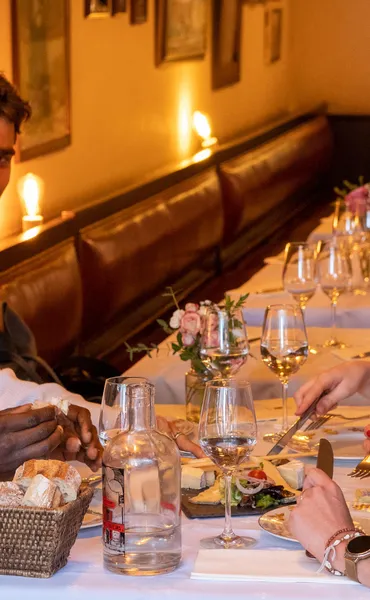 Close-up of people dining at a well-lit European restaurant, with bread, water, wine glasses, and cheese on table.