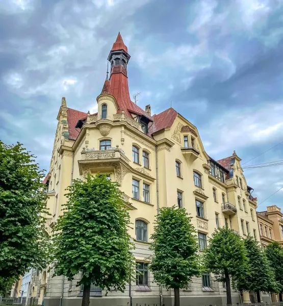 Edificio Art Nouveau ornamentado en Riga con fachada amarilla, techo rojo y torre, enmarcado por árboles verdes bajo cielo...