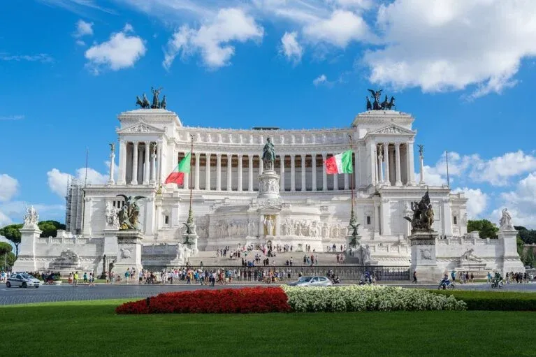 Gran Altar de la Patria de mármol blanco en Roma bajo un cielo azul con nubes. Banderas italianas a cada lado. Gente en la...