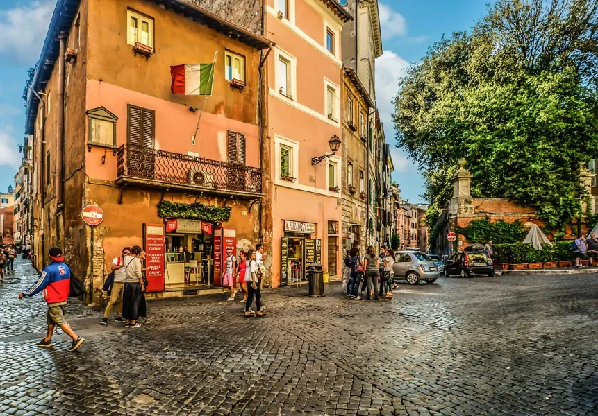 Calle adoquinada en Roma con edificios históricos, bandera italiana, personas caminando y coches aparcados bajo un cielo a...