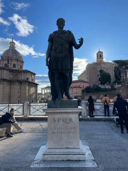 Bronzestatue von Julius Cäsar in Rom, mit Ruinen des Forum Romanum und einer Kuppelkirche unter blauem Himmel.