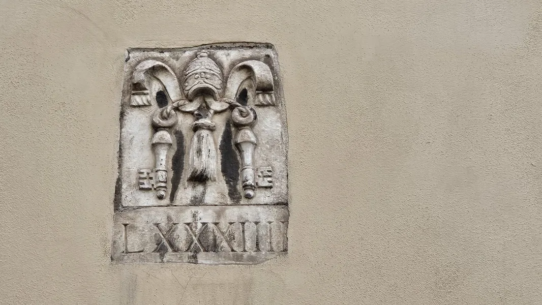 Kamienna płaskorzeźba na teksturowanej ścianie, przedstawiająca tiarę papieską, skrzyżowane klucze, frędzle i rzymskie cyf...