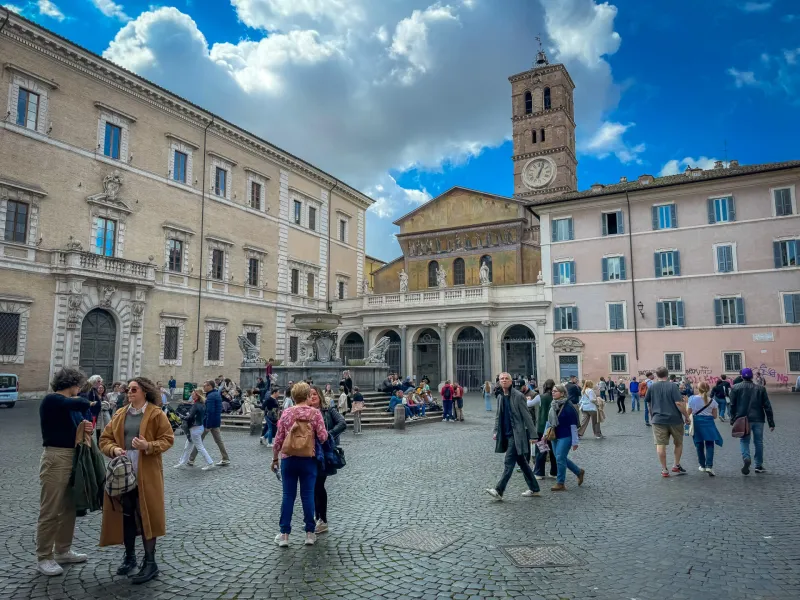 Brukowana Piazza Santa Maria w Trastevere, Rzym, z bazyliką, dzwonnicą, fontanną i spacerującymi ludźmi.