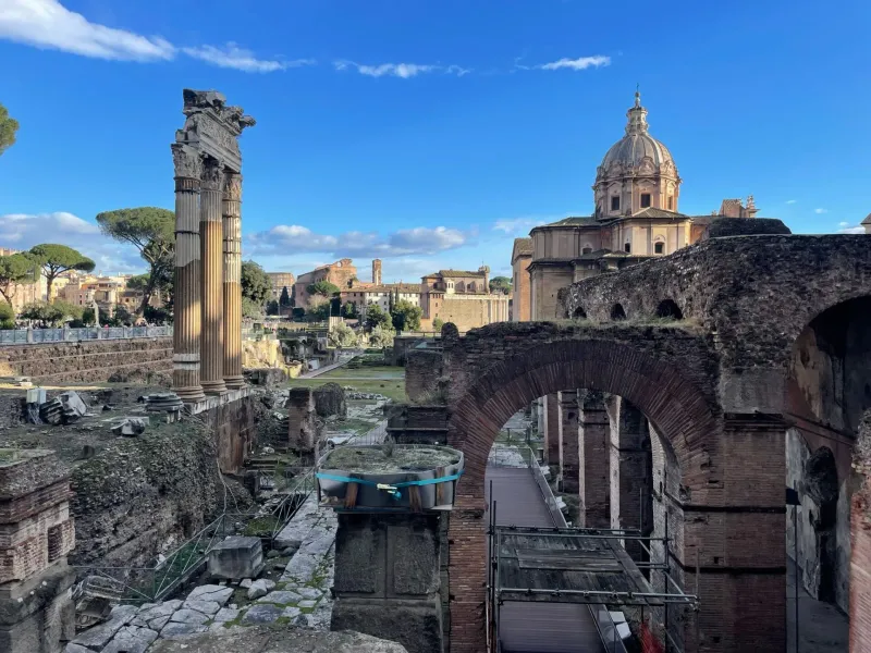 Ruinen des Forum Romanum mit Säulen des Saturn-Tempels, weiteren Tempeln und Besuchern unter blauem Himmel.