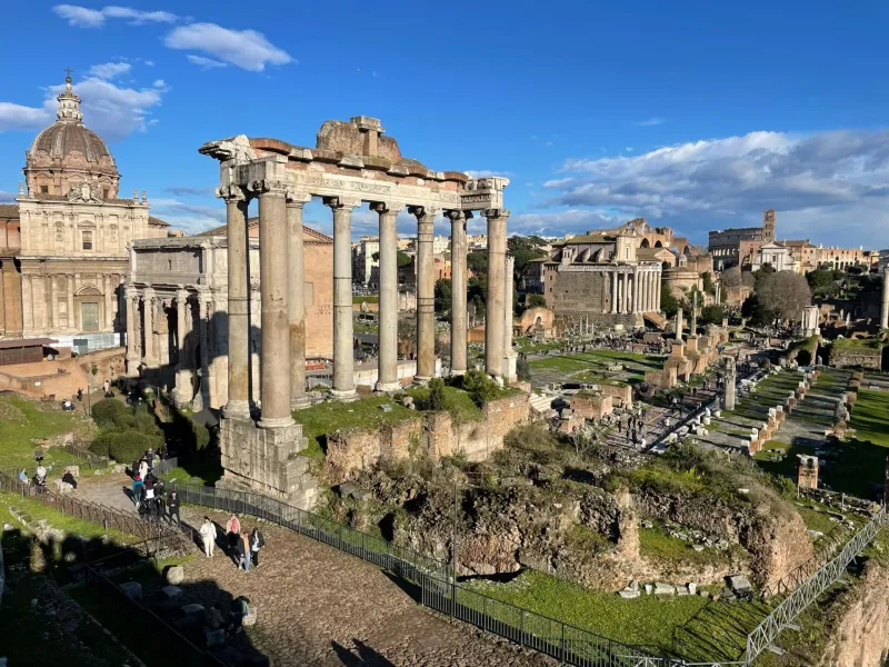 Ruinen des Forum Romanum mit Säulen des Saturn-Tempels, weiteren Tempeln und Besuchern unter blauem Himmel.