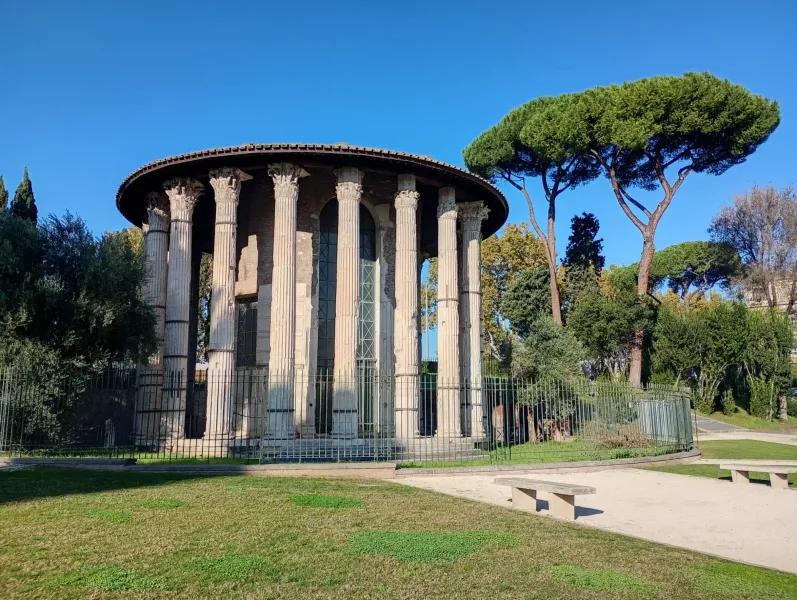 Ancient round Temple of Hercules Victor in Rome featuring Corinthian columns, surrounded by trees and a metal fence.
