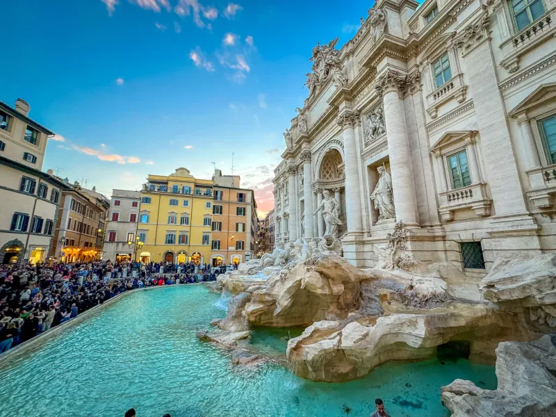 Abarrotada Fontana di Trevi en Roma al atardecer, con esculturas barrocas, agua turquesa y edificios históricos bajo un ci...